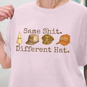 以下が含まれることがあります： 「Same Shit. Different Hat.」のテキストと、KKKのフード、軍用キャップ、「Make America Great Again」の帽子のイラストが描かれた、薄いピンク色のTシャツ。Tシャツはクルーネックです。