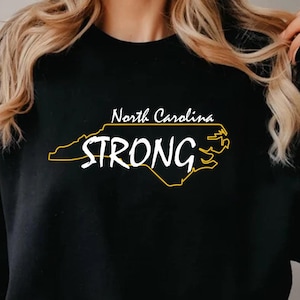 以下が含まれることがあります： 黒のトレーナーに、ノースカロライナ州の黄色のアウトラインと「North Carolina STRONG」という白い文字が書かれています。