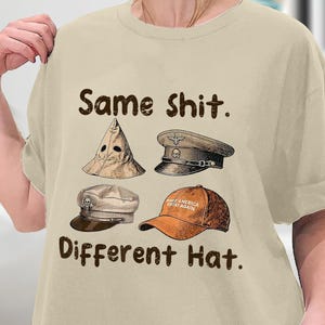 May include: Beige t-shirt with the text "Same Shit. Different Hat." and illustrations of four different hats. The hats include a pointed hood, a military cap, a flat cap, and a baseball cap.