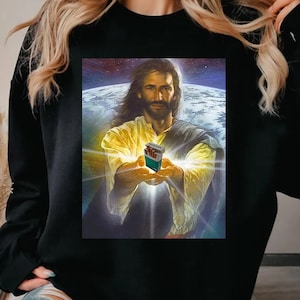 May include: Black sweatshirt featuring a graphic of Jesus holding a pack of cigarettes, set against a backdrop of Earth and space. The image has a religious theme. The brand name "CaHo Gifts" is visible.