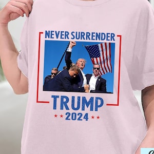 May include: Pink t-shirt with a graphic of a man holding a flag and the text "Never Surrender Trump 2024".
