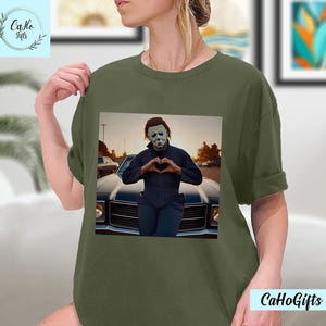 K&ouml;nnte beinhalten: Ein gr&uuml;nes T-Shirt mit einem Motiv von Michael Myers aus der Halloween-Filmreihe. Er steht vor einem Auto und formt mit seinen H&auml;nden ein Herz. Der Text "CaHoGifts" ist in der rechten unteren Ecke des Bildes zu sehen.