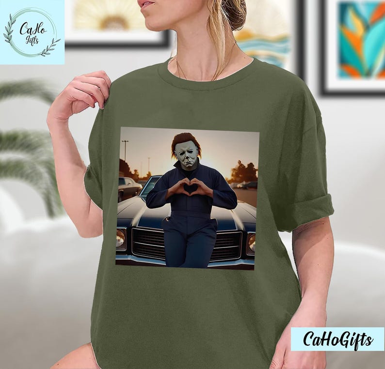 K&ouml;nnte beinhalten: Ein gr&uuml;nes T-Shirt mit einem Motiv von Michael Myers aus der Halloween-Filmreihe. Er steht vor einem Auto und formt mit seinen H&auml;nden ein Herz. Der Text "CaHoGifts" ist in der rechten unteren Ecke des Bildes zu sehen.
