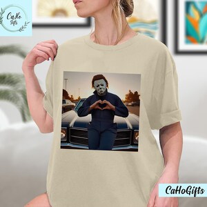 K&ouml;nnte beinhalten: Ein beiges T-Shirt mit einem Grafikdruck von Michael Myers aus der Halloween-Franchise. Die Grafik zeigt Michael Myers in einem blauen Overall, der mit seinen H&auml;nden ein Herz formt und vor einem Oldtimer steht.