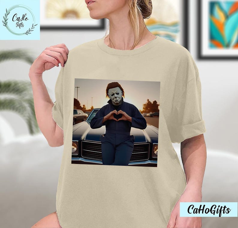 K&ouml;nnte beinhalten: Ein beiges T-Shirt mit einem Grafikdruck von Michael Myers aus der Halloween-Franchise. Die Grafik zeigt Michael Myers in einem blauen Overall, der mit seinen H&auml;nden ein Herz formt und vor einem Oldtimer steht.