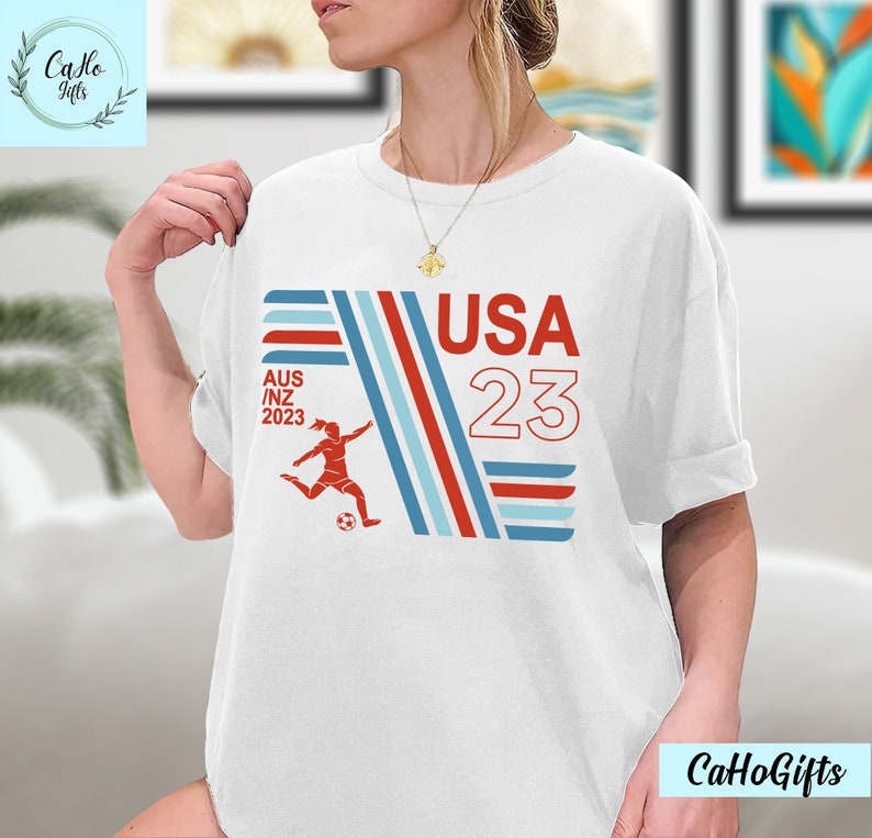 May include: White t-shirt with a red, white, and blue graphic design. The graphic features a female soccer player kicking a ball, the text "USA 23", and the text "AUS/NZ 2023".