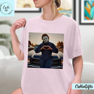 K&ouml;nnte beinhalten: Ein pinkfarbenes T-Shirt mit einem Motiv von Michael Myers aus der Halloween-Reihe. Er tr&auml;gt eine wei&szlig;e Maske und formt mit seinen H&auml;nden ein Herz. Er steht vor einem schwarzen Auto.