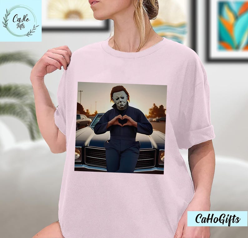 K&ouml;nnte beinhalten: Ein pinkfarbenes T-Shirt mit einem Motiv von Michael Myers aus der Halloween-Reihe. Er tr&auml;gt eine wei&szlig;e Maske und formt mit seinen H&auml;nden ein Herz. Er steht vor einem schwarzen Auto.
