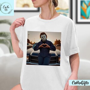 K&ouml;nnte beinhalten: Wei&szlig;es T-Shirt mit einem Motiv von Michael Myers aus der Halloween-Franchise, der vor einem Oldtimer ein Herz mit seinen H&auml;nden formt.