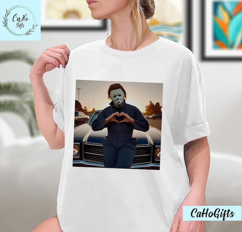 K&ouml;nnte beinhalten: Wei&szlig;es T-Shirt mit einem Motiv von Michael Myers aus der Halloween-Franchise, der vor einem Oldtimer ein Herz mit seinen H&auml;nden formt.
