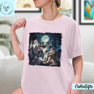 May include: A pink t-shirt with a graphic print of a group of werewolves howling at the moon. The werewolves are in a dark forest setting with a full moon in the background.