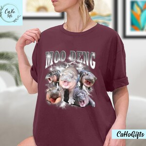 K&ouml;nnte beinhalten: Ein maroonfarbenes T-Shirt mit dem Text "MOO DENG" in silbernen Buchstaben mit einem Blitz-Design. Das Shirt zeigt eine Grafik mit f&uuml;nf Baby-Fluss Pferden.