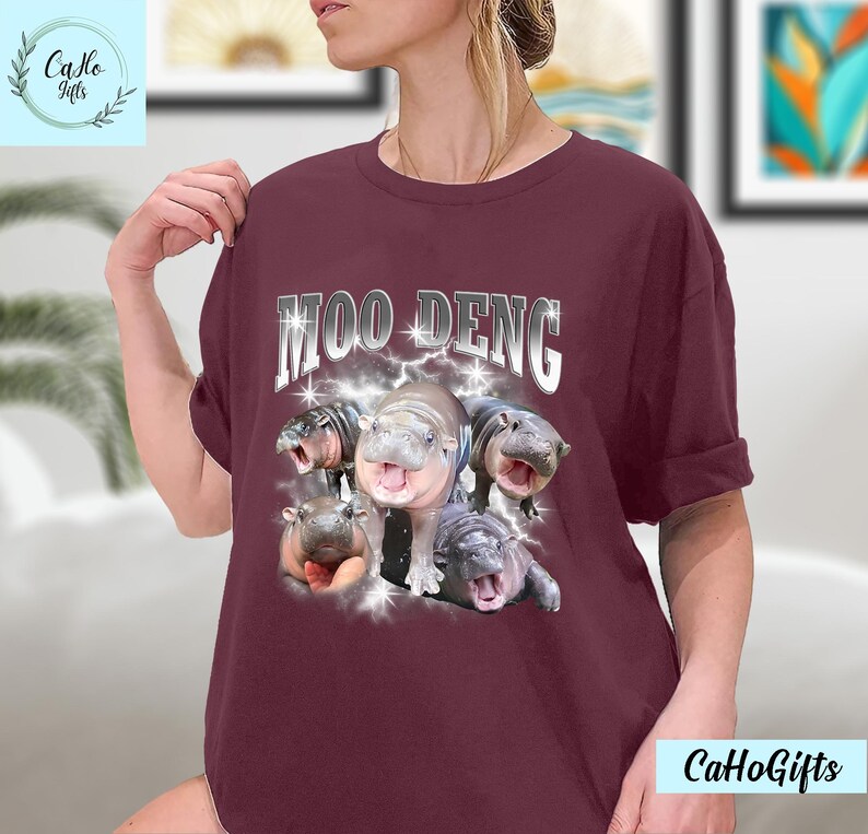 K&ouml;nnte beinhalten: Ein maroonfarbenes T-Shirt mit dem Text "MOO DENG" in silbernen Buchstaben mit einem Blitz-Design. Das Shirt zeigt eine Grafik mit f&uuml;nf Baby-Fluss Pferden.