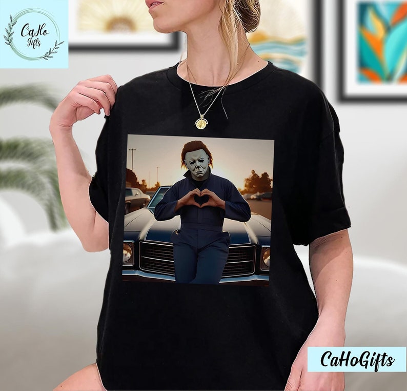 K&ouml;nnte beinhalten: Ein schwarzes T-Shirt mit einem Grafikdruck von Michael Myers aus der Halloween-Filmreihe. Er steht vor einem Auto und formt mit seinen H&auml;nden ein Herz.