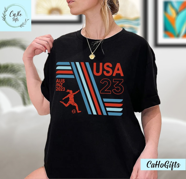 May include: Black t-shirt with a red, white, and blue graphic design. The graphic features a soccer player kicking a ball, the text "USA 23", and the text "AUS/NZ 2023".