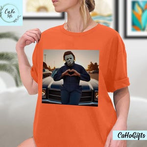 K&ouml;nnte beinhalten: Orangefarbenes T-Shirt mit einem Grafikdruck von Michael Myers aus der Halloween-Filmreihe. Er steht vor einem Auto und formt mit seinen H&auml;nden ein Herz.