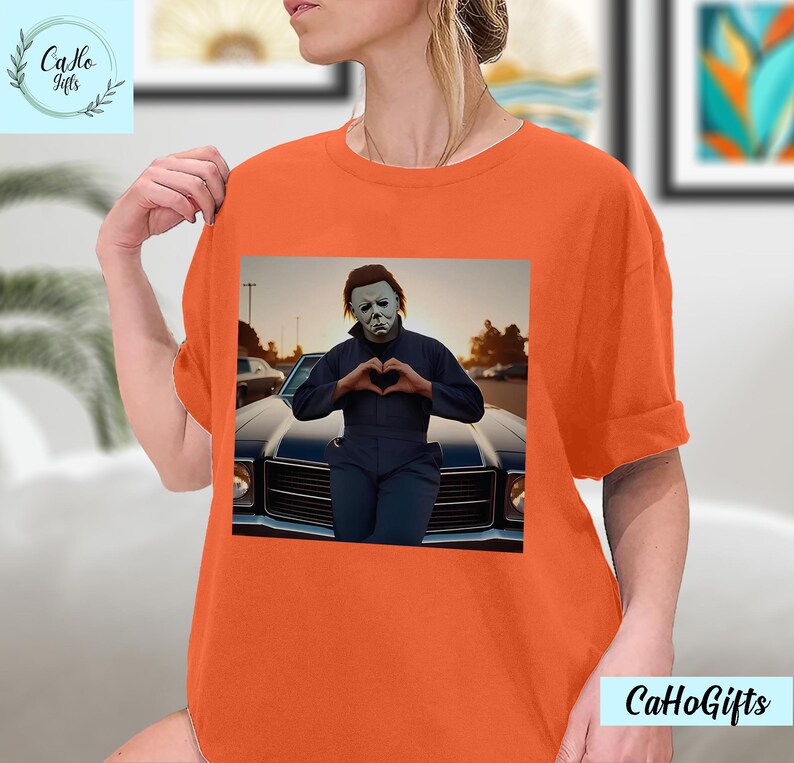K&ouml;nnte beinhalten: Orangefarbenes T-Shirt mit einem Grafikdruck von Michael Myers aus der Halloween-Filmreihe. Er steht vor einem Auto und formt mit seinen H&auml;nden ein Herz.