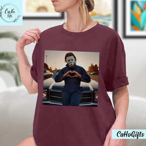 K&ouml;nnte beinhalten: Ein maroonfarbenes T-Shirt mit einem Motiv von Michael Myers aus der Halloween-Reihe. Er tr&auml;gt eine wei&szlig;e Maske und formt mit seinen H&auml;nden ein Herz. Er steht vor einem Oldtimer.