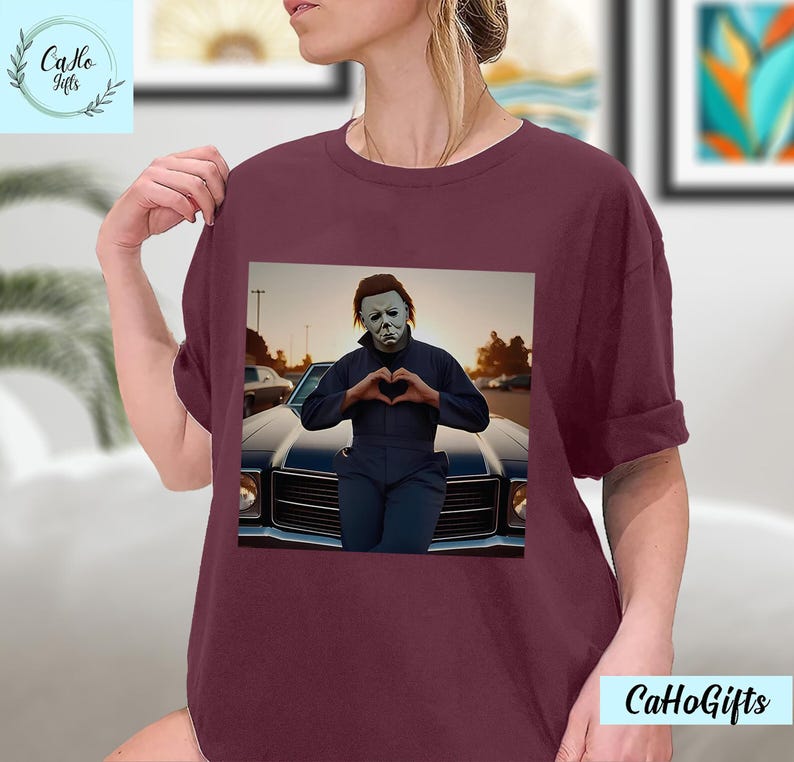 K&ouml;nnte beinhalten: Ein maroonfarbenes T-Shirt mit einem Motiv von Michael Myers aus der Halloween-Reihe. Er tr&auml;gt eine wei&szlig;e Maske und formt mit seinen H&auml;nden ein Herz. Er steht vor einem Oldtimer.
