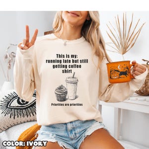 Puede incluir: Sudadera de color marfil con el texto "This is my: running late but still getting coffee shirt." Un gráfico de cupcake y taza de café está debajo. Se sostiene una taza naranja de Halloween con un gato negro y una calabaza.