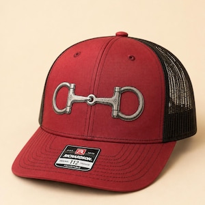 May include: A maroon trucker hat featuring a black mesh back and a silver-toned metal horse bit emblem centered on the front panel. The brim displays a black sticker with white text reading Richardson Original 112 Trucker Adjustable.