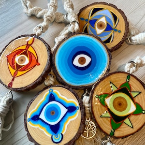 May include: Five wooden round ornaments with blue and white evil eye designs painted on them. The ornaments are hanging from a macrame cord with tassels.