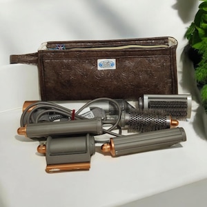 May include: A set of four hair styling tools with a brown zippered pouch. The tools are gray and rose gold in color and have a cord. The pouch has a floral pattern and the text "Trish McEvoy" on it.