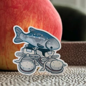 May include: A sticker of a blue fish wearing white trainers is placed in front of a red apple. The apple has a red and yellow gradient. The background is blurred, with a glimpse of a window and a dark green object.