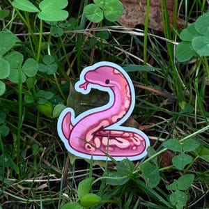 May include: A pink snake sticker with a white border, resting on a bed of green grass and clover. The sticker features a stylized snake design with a light pink body and darker pink markings.