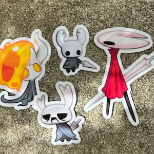 May include: Four character stickers from the video game Hollow Knight. The stickers show cartoon-style characters in white, orange, red, and black. The stickers are placed on a textured, neutral-toned surface, likely a table or similar.