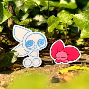 May include: Two stickers are displayed outdoors among green foliage. One is a white, cartoonish skull with blue accents. The other is a pink, flower-like character with a grumpy expression. The stickers are on a stone surface.