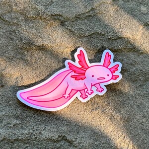 K&ouml;nnte beinhalten: Ein rosa Axolotl-Aufkleber mit wei&szlig;em Rand. Der Cartoon-Axolotl hat rosa Kiemen, einen langen Schwanz und kleine Beine. Der Aufkleber befindet sich auf einer strukturierten, hellbraunen Oberfl&auml;che.
