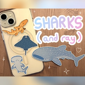 a small, cute pastel whale shark sticker, hammerhead shark sticker, stingray sticker, and epaulette/walking shark sticker sitting on a wooden table and on the back of an iphone