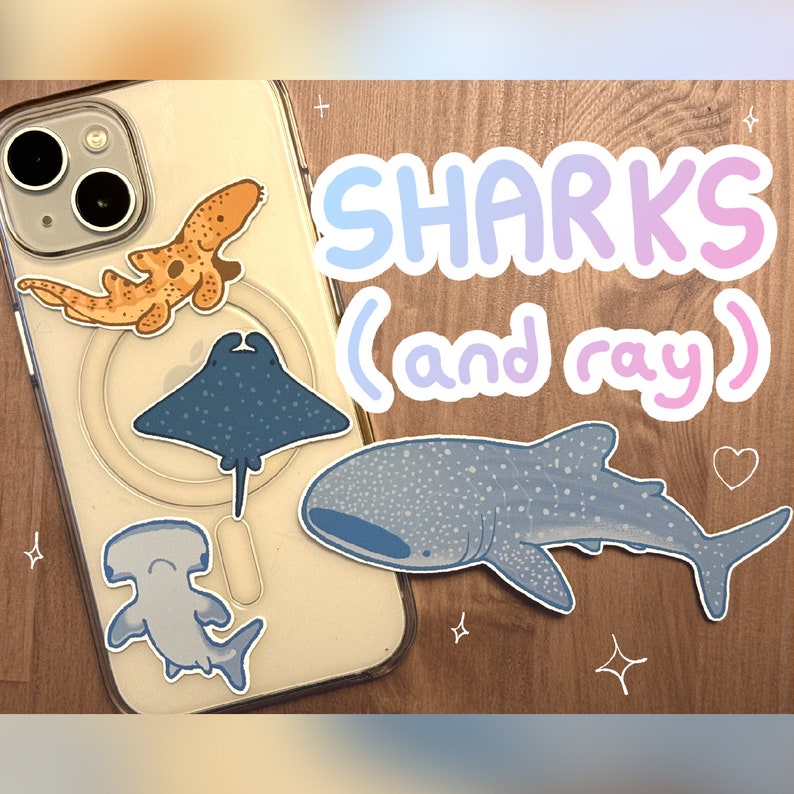 a small, cute pastel whale shark sticker, hammerhead shark sticker, stingray sticker, and epaulette/walking shark sticker sitting on a wooden table and on the back of an iphone