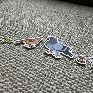 May include: A set of five stickers featuring a banana peel, a sparrow, a pigeon, an apple core, and a cup of coffee. The stickers are on a light brown textured surface.