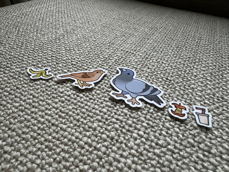 May include: A set of five stickers featuring a banana peel, a sparrow, a pigeon, an apple core, and a cup of coffee. The stickers are on a light brown textured surface.