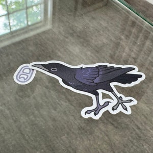 May include: A black crow sticker with a white outline, holding a silver ring in its beak. The sticker is on a glass surface.