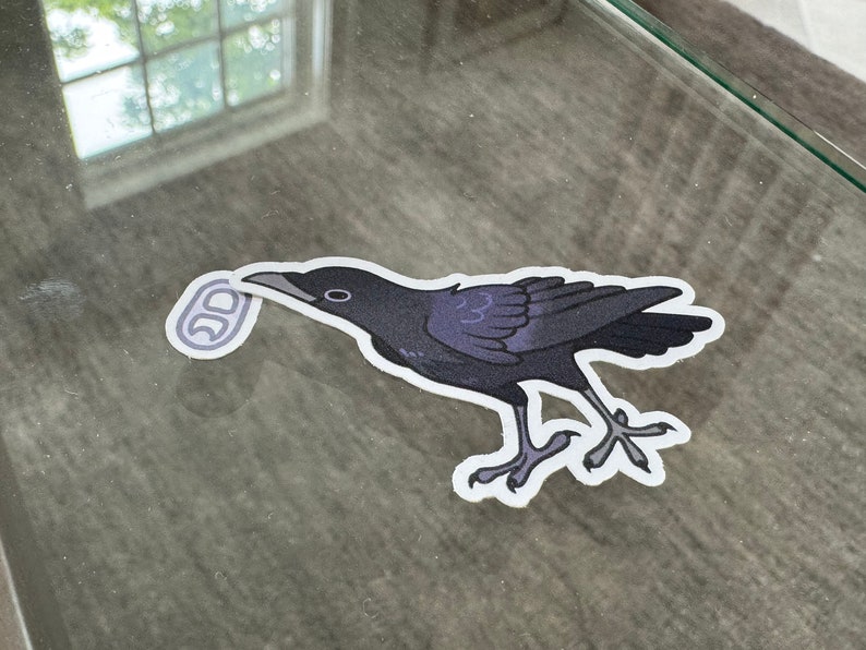 May include: A black crow sticker with a white outline, holding a silver ring in its beak. The sticker is on a glass surface.