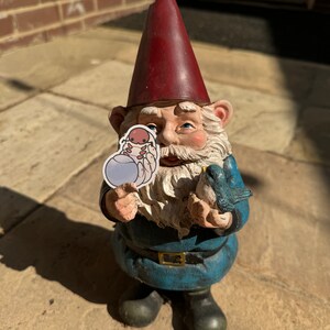 May include: A garden gnome with a red hat and blue clothing is holding a small blue orb with a cartoon creature inside. The gnome is also holding a blue bird in its other hand.