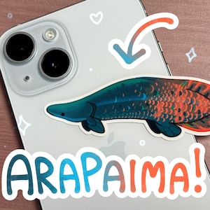 May include: A sticker of a colorful Arapaima fish on a white iPhone case. The sticker has a blue, green, and red color scheme. The text "ARAPAIMA!" is written in a colorful font below the fish.