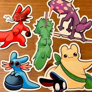 Op de afbeelding: Een set kleurrijke cartoon dieren stickers. De stickers bevatten een rode vos, een groen wezen, een paars-zwarte hagedis, een geel wezen met een groene sjerp, een blauwe axolotl en een zwart-rood wezen.