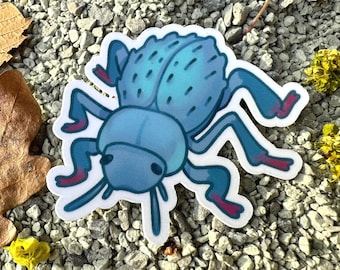 May include: A die-cut sticker featuring a stylised blue beetle with a rounded body, textured markings on its back, and distinct reddish-pink accents on the tips of its six legs. The illustration has a white border and a smooth, matte finish.