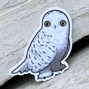 May include: A white snowy owl sticker with yellow eyes. The owl is facing forward with its wings tucked in. The sticker has a white background.