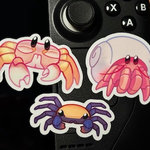 May include: Three cartoon crab stickers on a black background. The first crab is orange and pink with large eyes. The second crab is pink and white with a shell and large eyes. The third crab is blue and orange with large eyes.