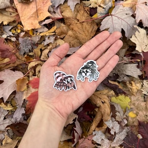 May include: Two small, colourful jumping spider stickers on a hand against a background of fallen autumn leaves. The stickers are about the size of a 10 pence coin.