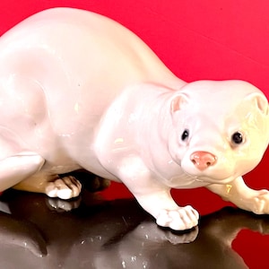 May include: A white ceramic ferret figurine with a glossy finish. The ferret is in a crawling position, with its paws extended. The figurine has black eyes and a pink nose, set against a red background.