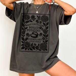 May include: Dark gray t-shirt with a black floral design and the text "WHAT EXCELLENT BOILED POTATOES EXEMPLARY VEGETABLE". The design includes two potatoes and decorative flowers.