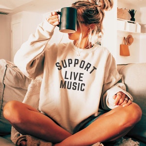 May include: A woman wearing a light beige sweatshirt with the text "SUPPORT LIVE MUSIC" printed on the front. She is sitting on a couch and holding a black mug.