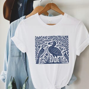 Puede incluir: Camiseta blanca con una ilustración azul marino de tres pájaros rodeados de follaje. El diseño está centrado en la camiseta. La camiseta está colgada en una percha de madera.