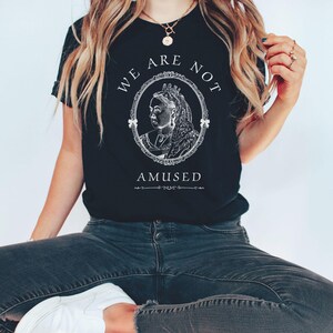 May include: A black t-shirt with a white graphic of a queen's portrait in a frame with the text "We Are Not Amused".